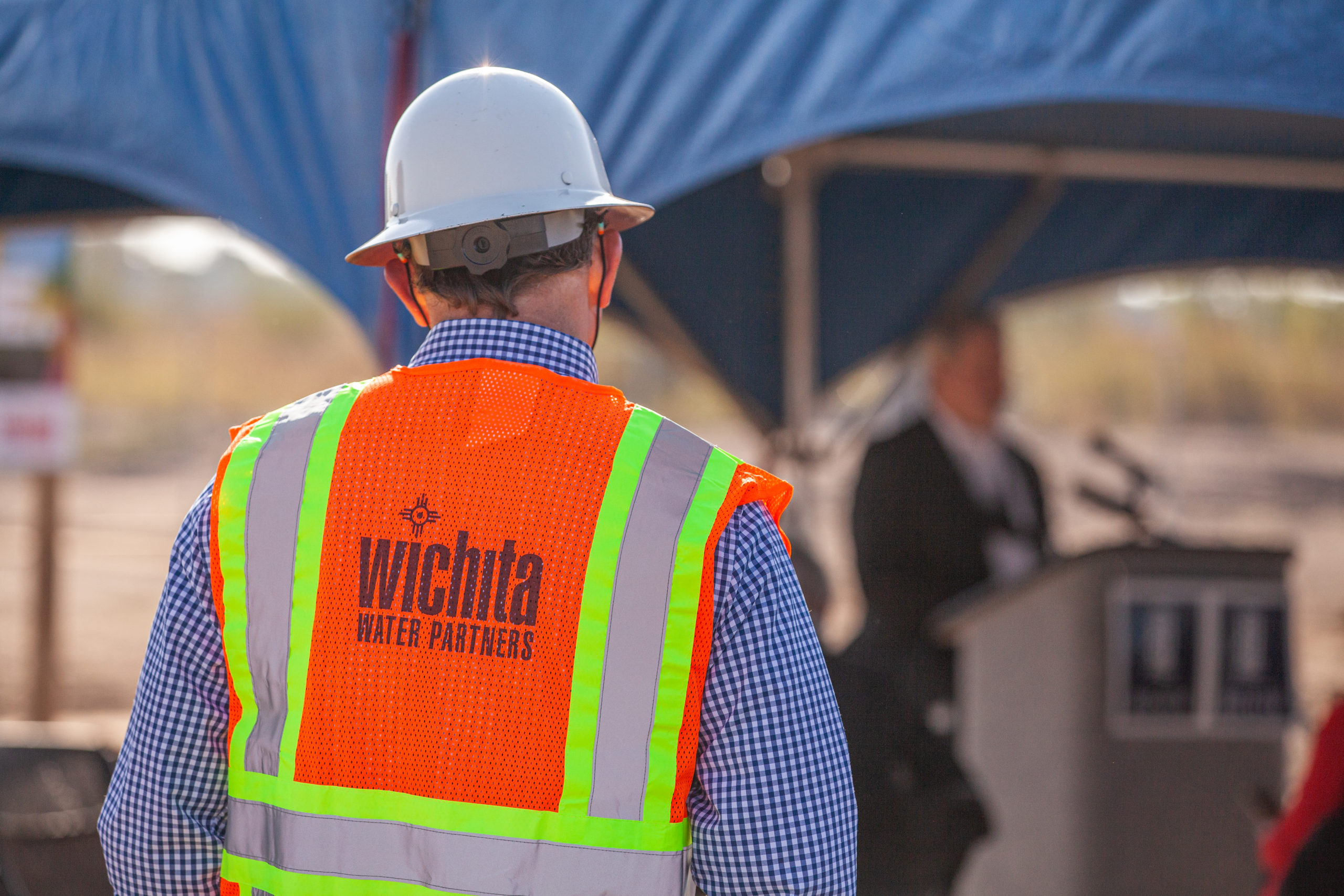 Photograph of the back of a man wearing Wichita Water Partners high visibility vest and hard hat at groundbreaking ceremony