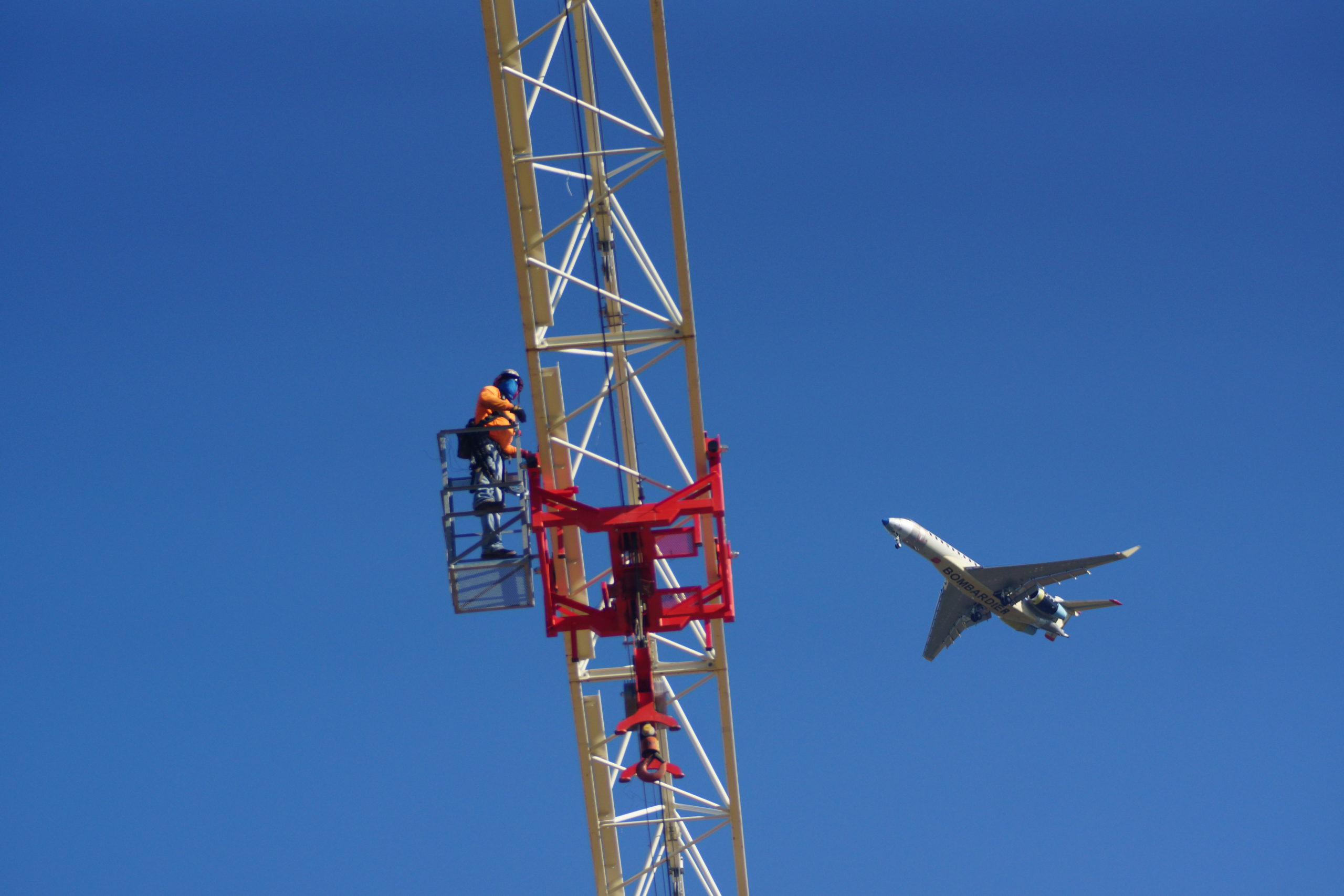 Image of tower crane boom with men dismantling it and an airplane flying in the sky in the background.