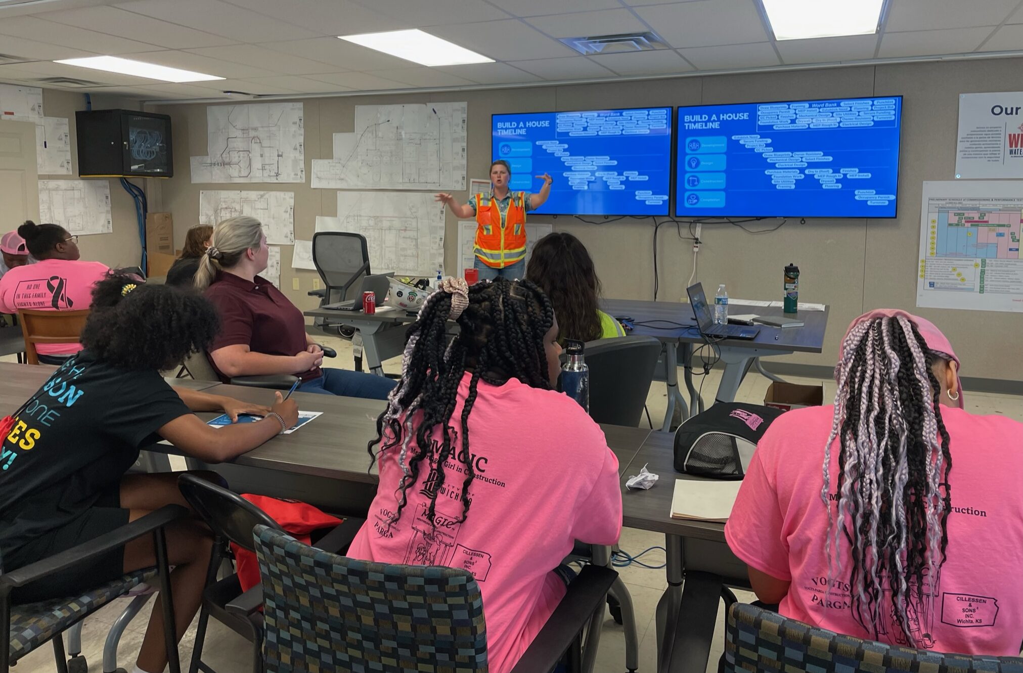 Photo shows a woman in a neon orange safety vest standing in front of two monitors presenting to a room full of teenage girls wearing pink shirts for the Mentoring A Girl in Construction Camp program