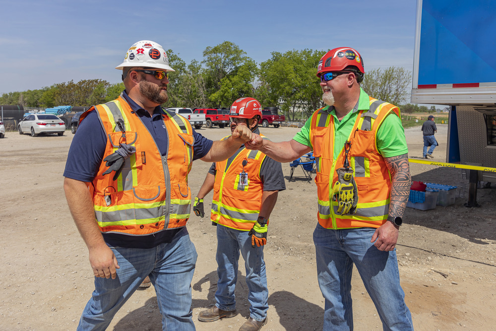 Two mean wearing orange safety vests and hard hats share a fist bump while another man in construction gear stands in the middle behind them